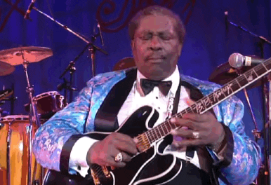 A grey-haired BB King is playing electric guitar in a blue paisley jacket and black bow-tie. He focuses hard while he passionately uses a technique called finger vibrato to get a bluesy sound out of the guitar.
