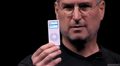 Steve Jobs holds up an iPod on stage. He is wearing his classic black turtle neck with thin metal round glasses.